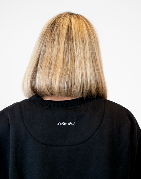 Rear view of woman wearing Ladies Black Relaxed Fleece Crew with "LUKE 18:1" text on back