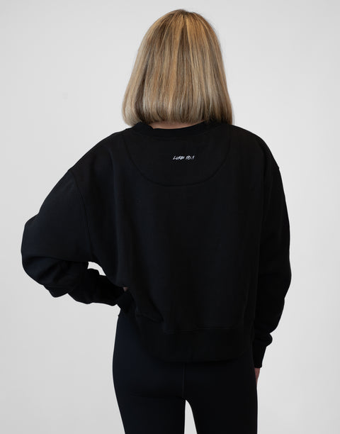 Rear view of woman wearing Ladies Black Relaxed Fleece Crew with "LUKE 18:1" text on back