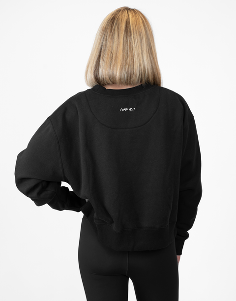 Rear view of woman wearing Ladies Black Relaxed Fleece Crew with "LUKE 18:1" text on back