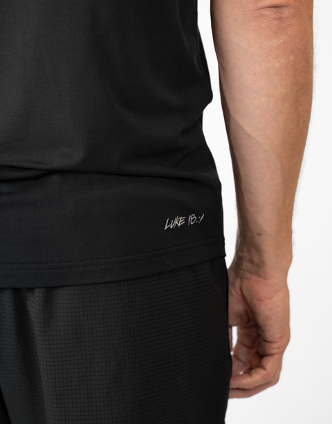 Rear view of man wearing Men's black Bamboo Performance Tee showing "LUKE 18:1" text on right back side near the bottom seam