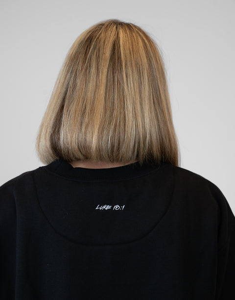 Rear view of woman wearing Ladies Black Relaxed Fleece Crew with "LUKE 18:1" text on back