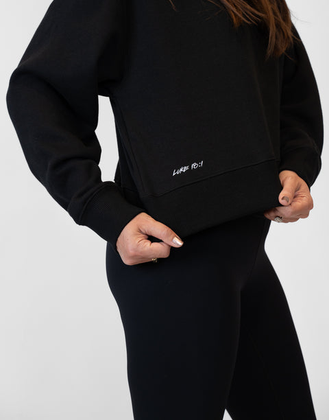 Person wearing Ladies Black Relaxed Fleece Crew with "LUKE 18:1" text on lower side
